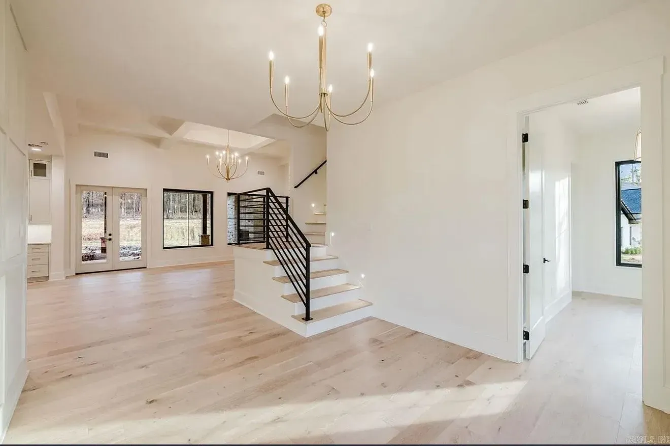 Bright, open foyer with light wood floors, a staircase with black railings, and two hanging chandeliers.