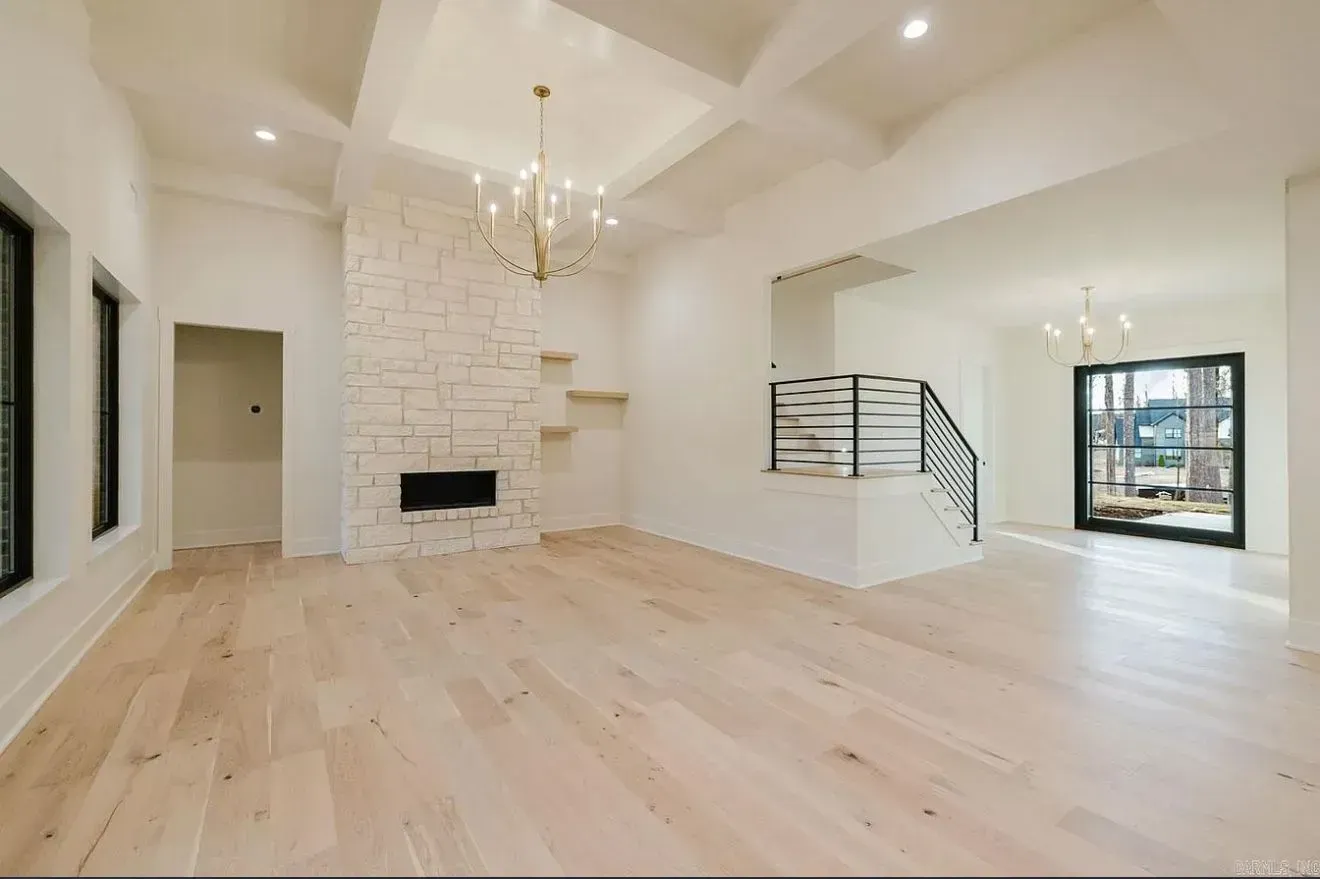 A spacious, bright room with light wood floors, a white stone fireplace, a coffered ceiling, and a staircase.