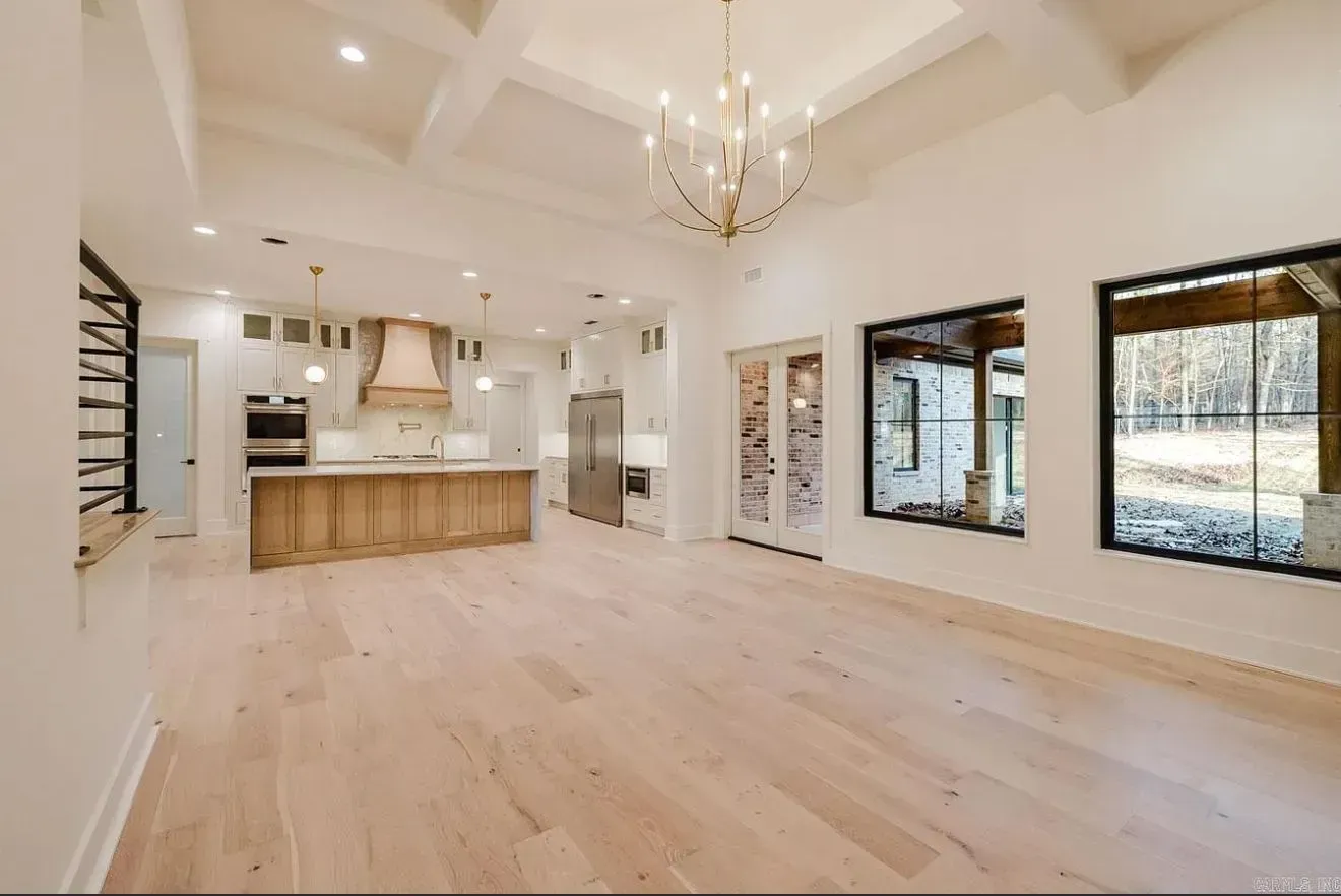A spacious, modern room with light wood floors, white walls, a coffered ceiling with a chandelier, and an open kitchen.