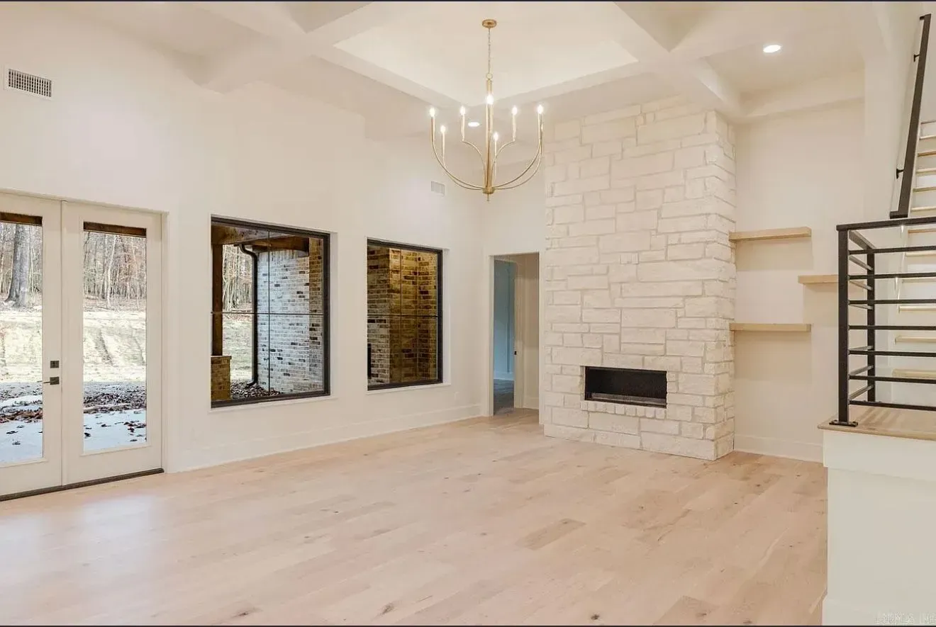A minimalist living room with white walls, light wood floors, a tall stone fireplace, and a gold modern chandelier.