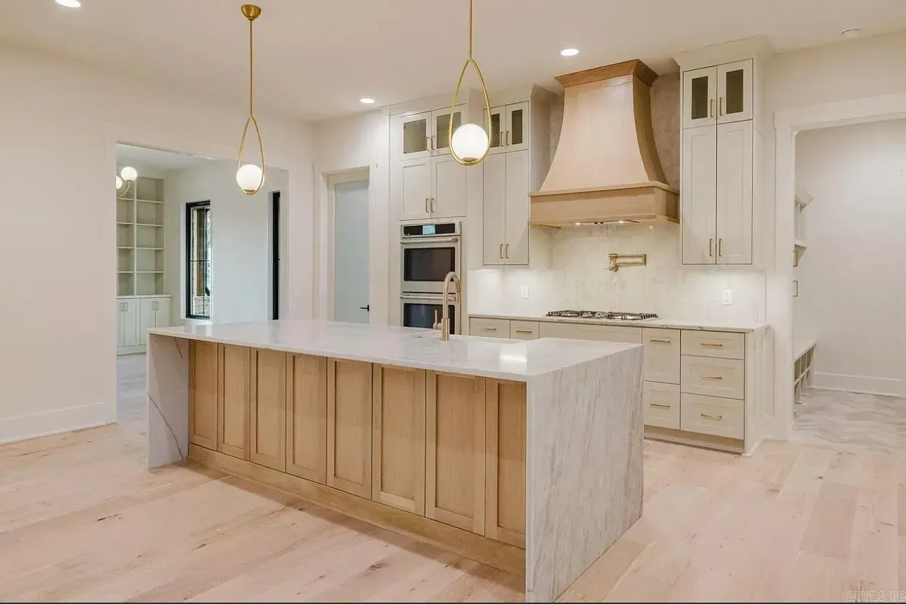 A bright, modern kitchen featuring a large marble-topped island with wood cabinetry, a range hood, and light wood floors.