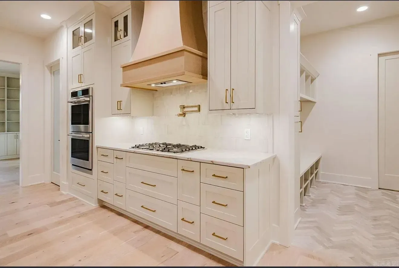A bright, modern kitchen featuring white cabinets, a wood range hood, a stainless double oven, and white marble counters.