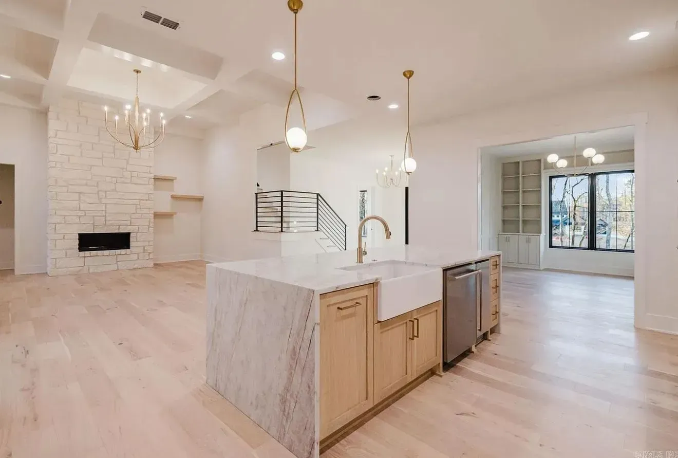 A bright kitchen and living area with light wood floors, a marble-topped island, a stone fireplace, and gold lighting.