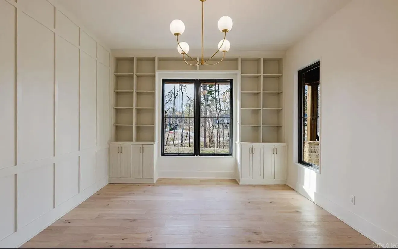 A bright, empty room with light wood floors, built-in shelving flanking a large window, and a modern globe chandelier.