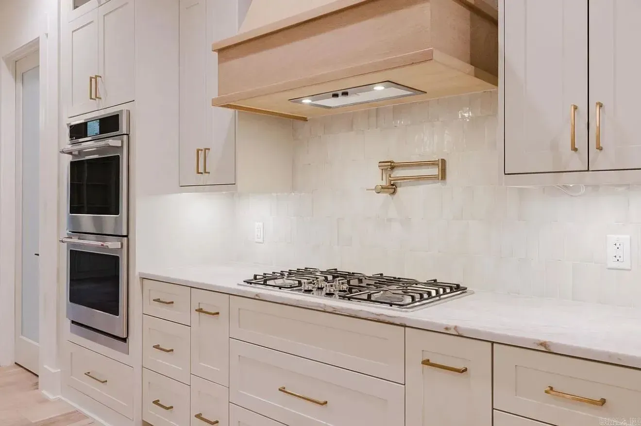 A modern kitchen featuring off-white cabinets, gold hardware, a gas cooktop, a wood range hood, and a wall-mounted faucet.