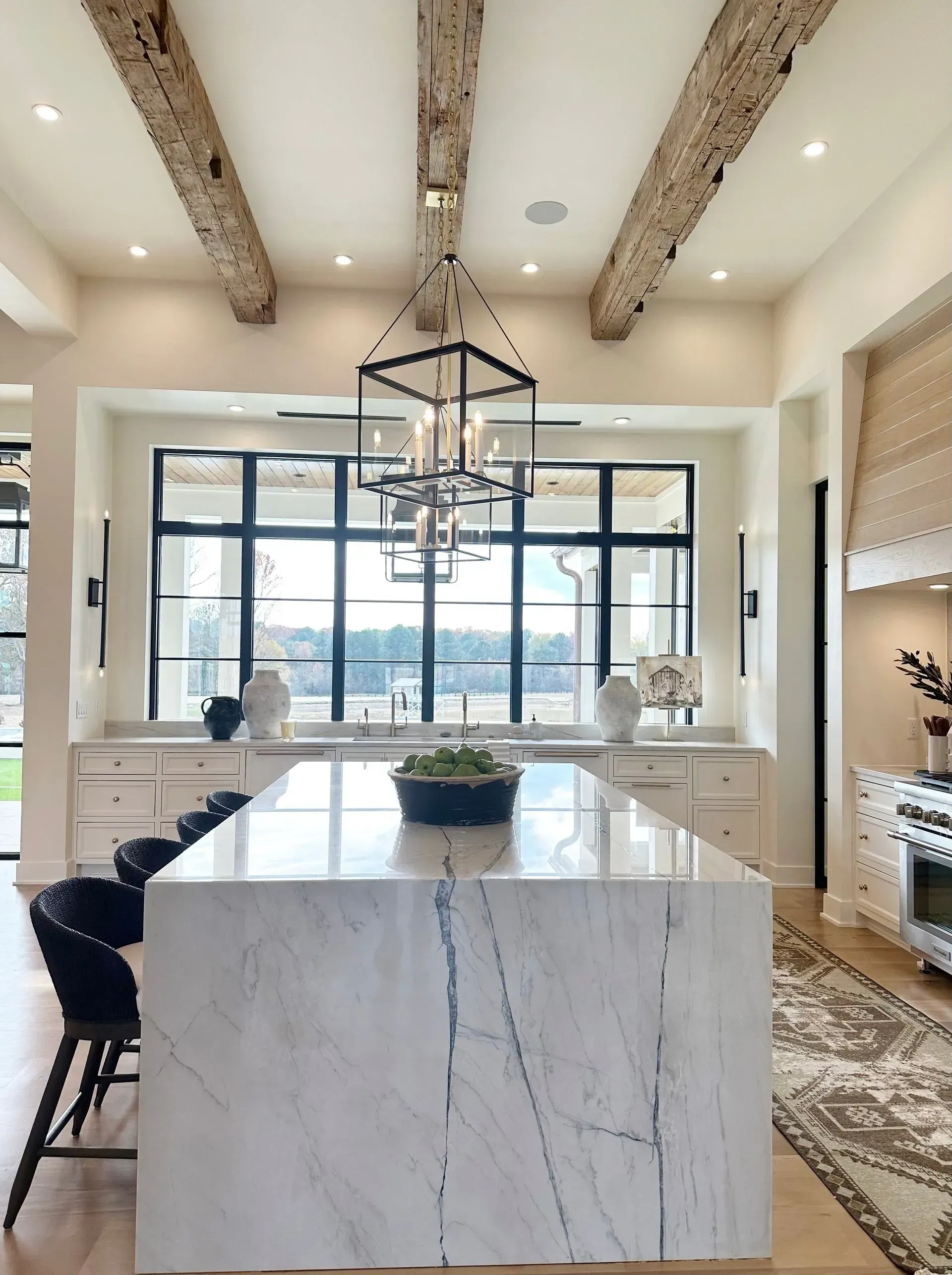 A kitchen with a large white marble waterfall island, black stools, rustic wood ceiling beams, and a windowed wall.