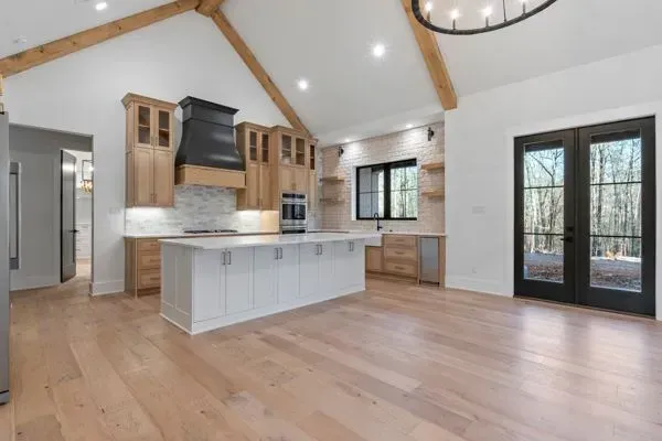 Modern kitchen with light wood cabinets, white island, black range hood, vaulted ceiling with beams, and black French doors.