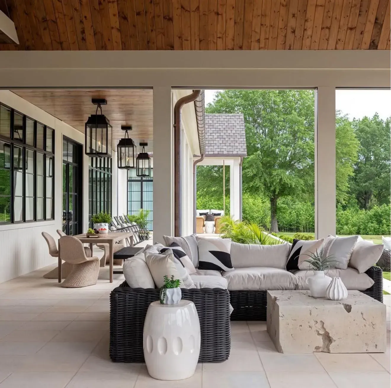 A covered outdoor patio featuring a gray wicker sectional sofa, a stone coffee table, and a dining area with pendant lights.