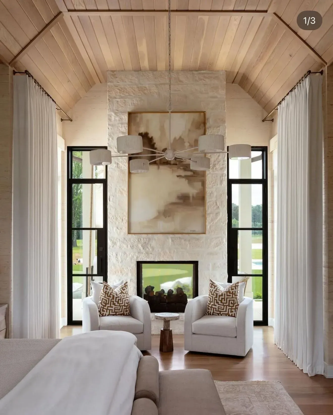 A bedroom with a vaulted wooden ceiling, a stone fireplace with a painting above it, and two chairs facing each other.
