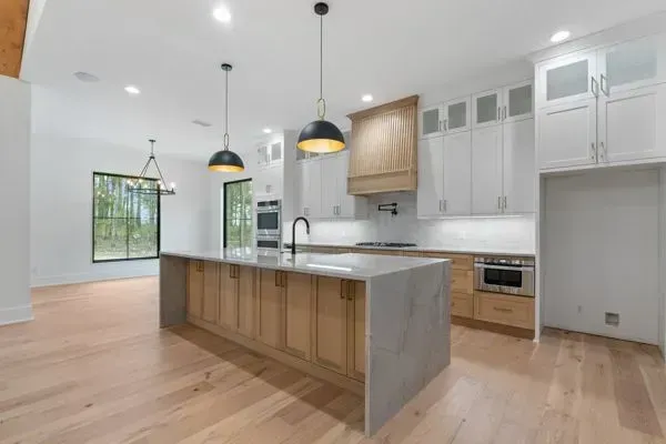 Modern kitchen with a gray waterfall island, light wood cabinetry, white upper cabinets, and a vent hood, and wood floors.