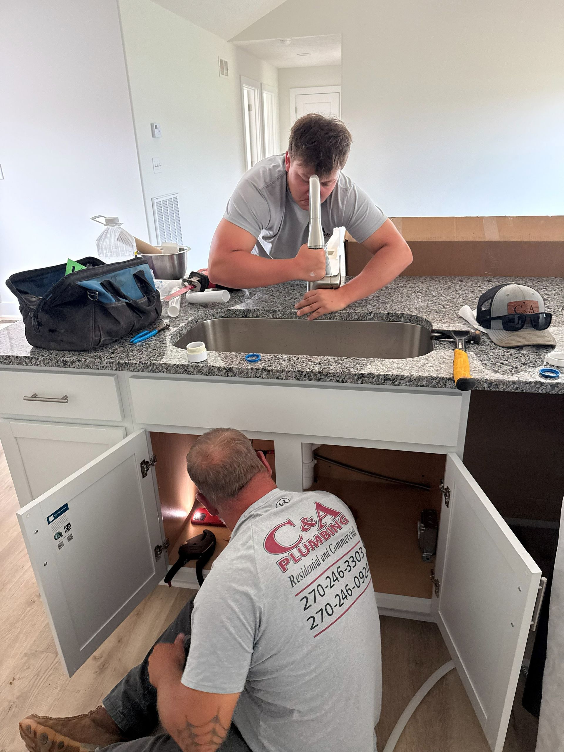 Two workers installing a kitchen sink and plumbing under a white countertop.