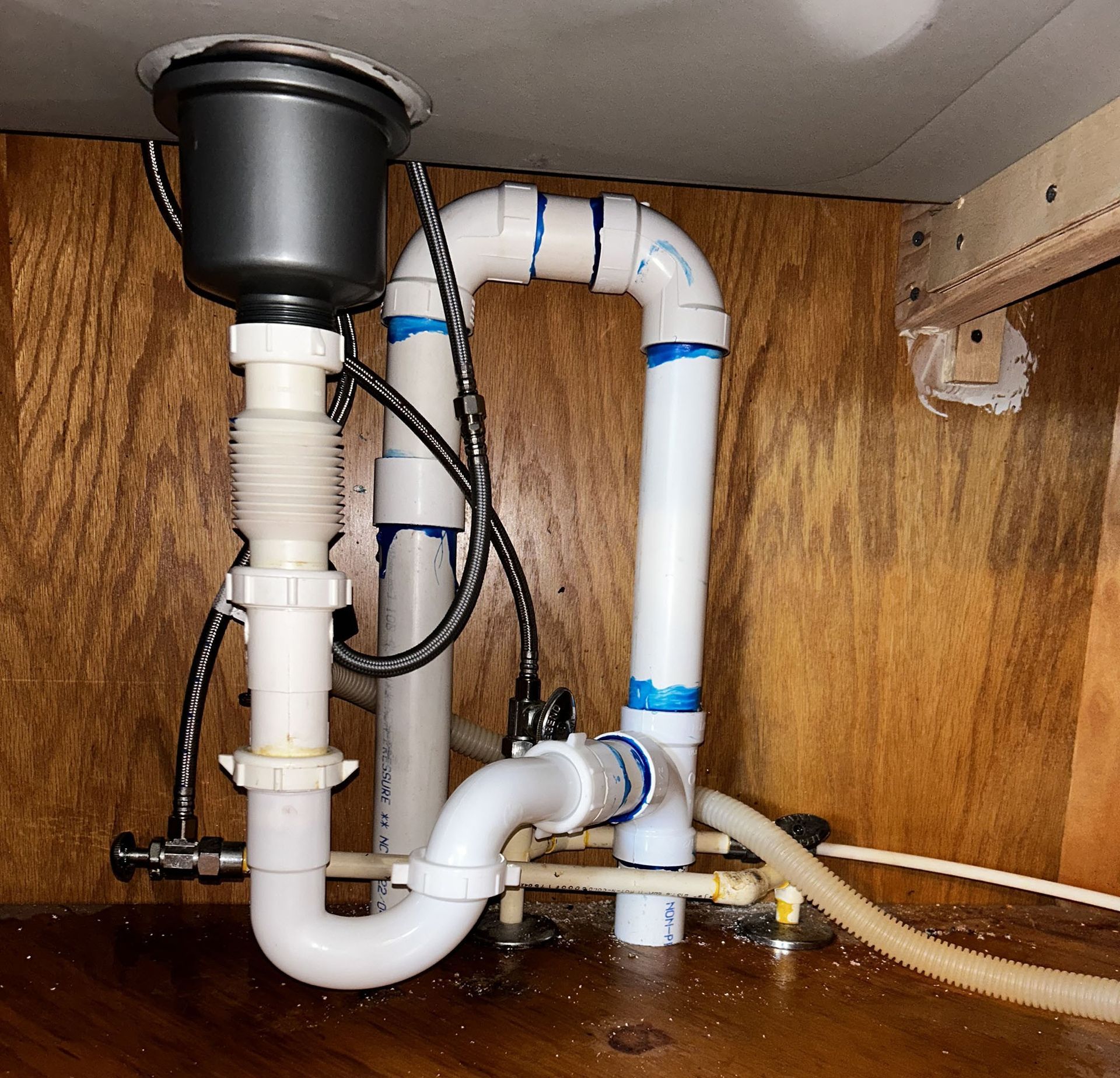 Under-sink plumbing with white PVC pipes, a black disposal, and a damp cabinet floor.