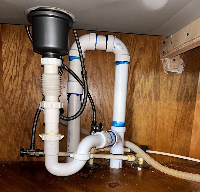 Under-sink plumbing with white PVC pipes, black disposal unit, and water on the cabinet floor.