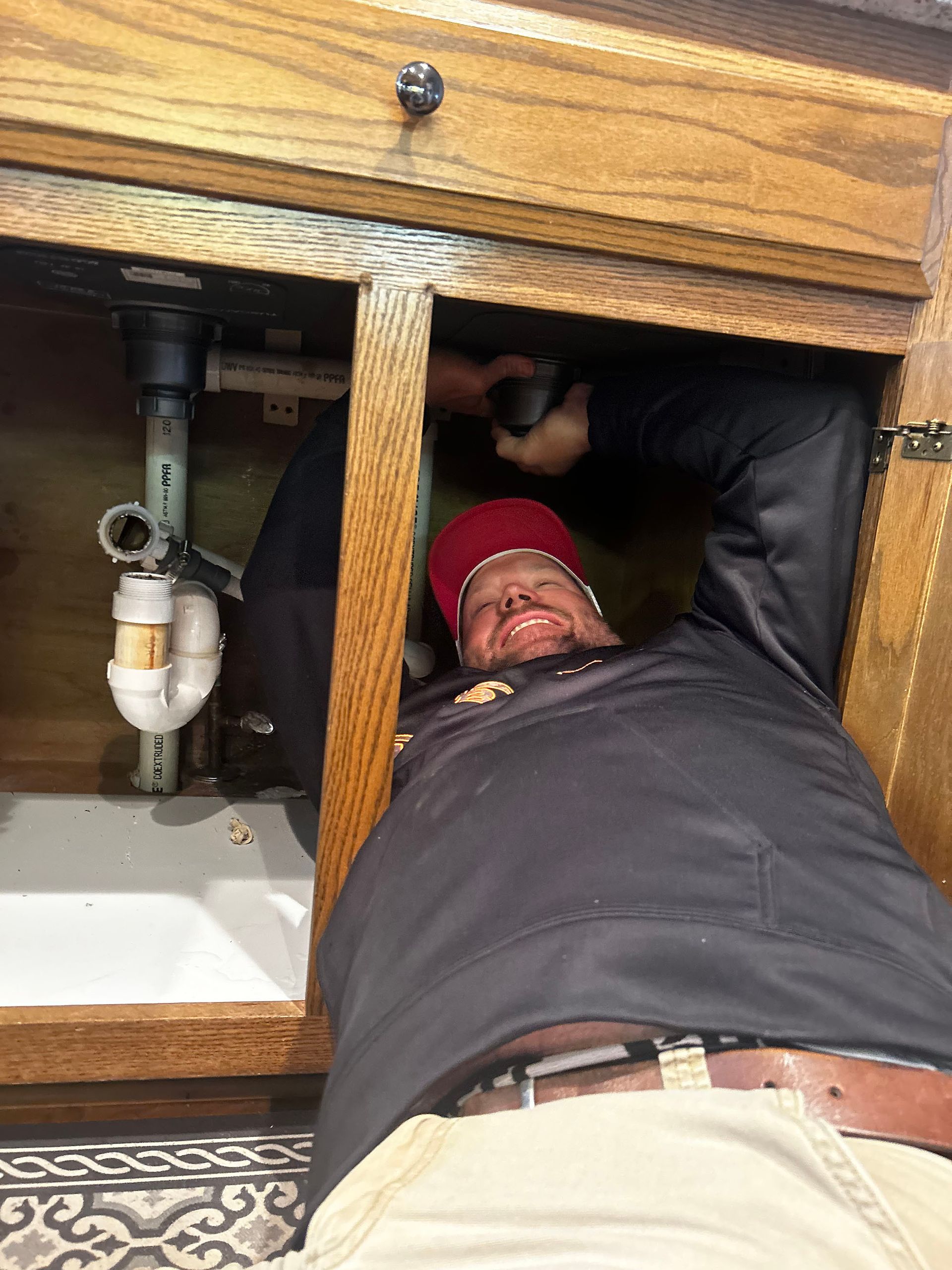 Plumber repairing under-sink pipes in a wooden cabinet, using a tool while lying on the floor.