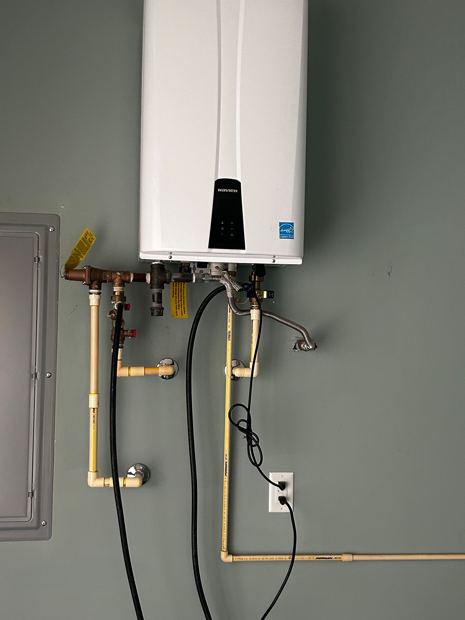 Wall-mounted water heater with exposed gas and water pipes in a utility room, against a gray wall.