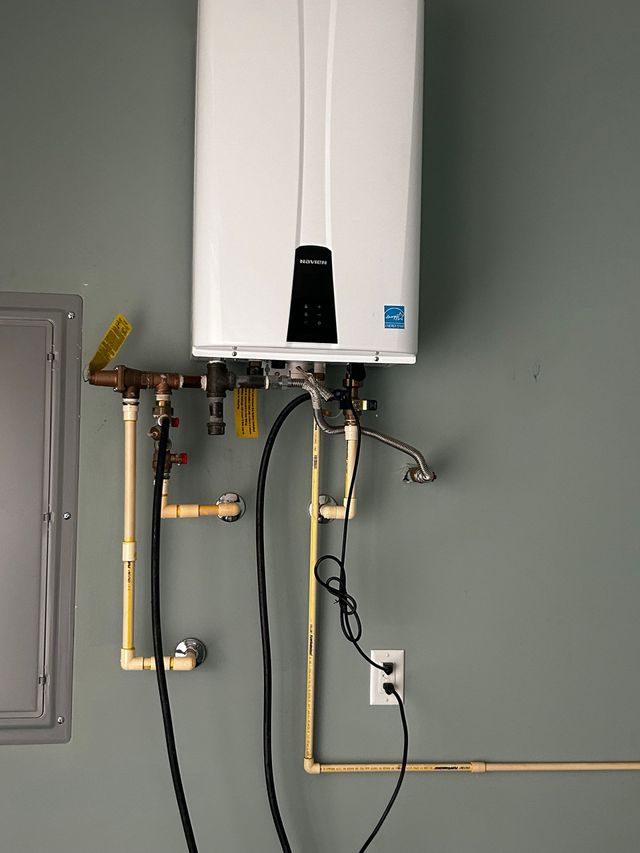 Wall-mounted water heater with exposed pipes and hoses against a gray utility room wall.