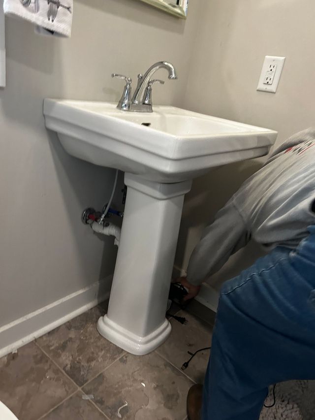 Bathroom pedestal sink beside a person in blue pants, with plumbing visible underneath.