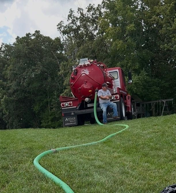 Person on a red vacuum truck with a green hose on a grassy slope near trees