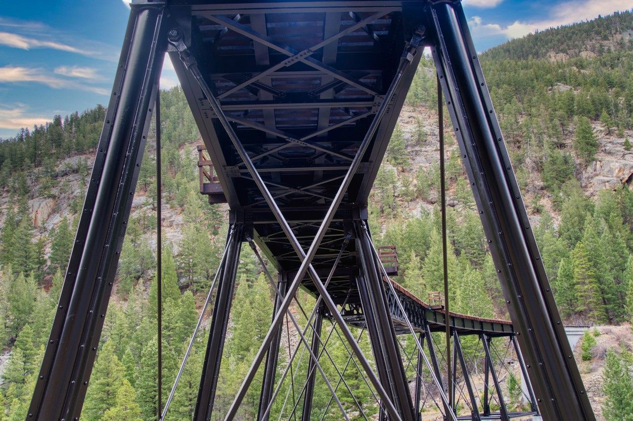 Underside of a black steel bridge spanning a rocky green canyon under a cloudy sky