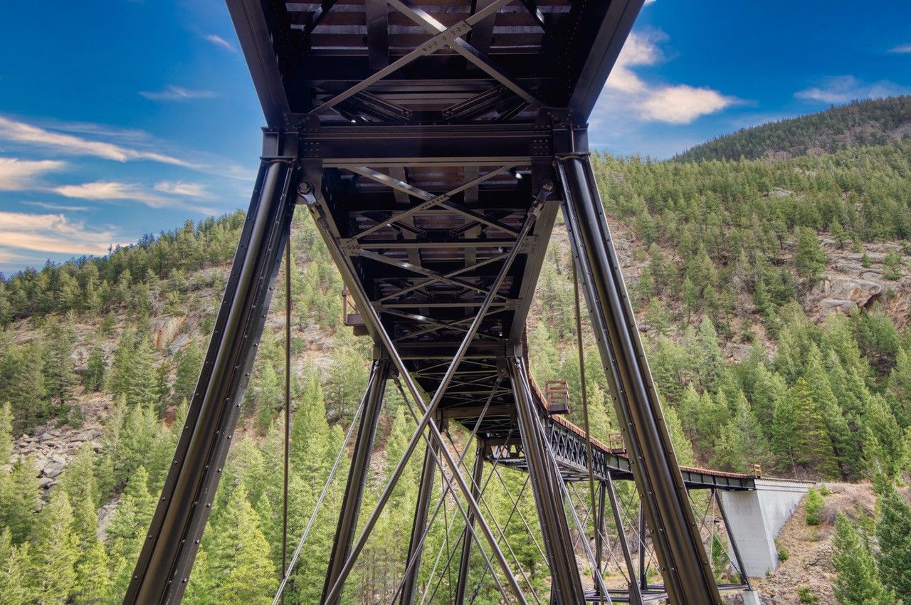 Underside of a steel bridge spanning a forested mountain valley under a blue sky.