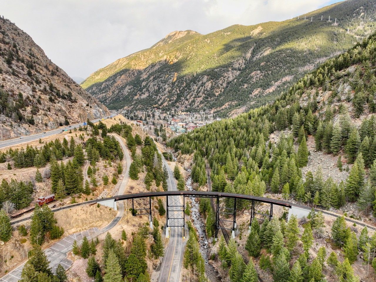 Curved bridge over a pine-filled mountain valley with rocky slopes and green peaks.