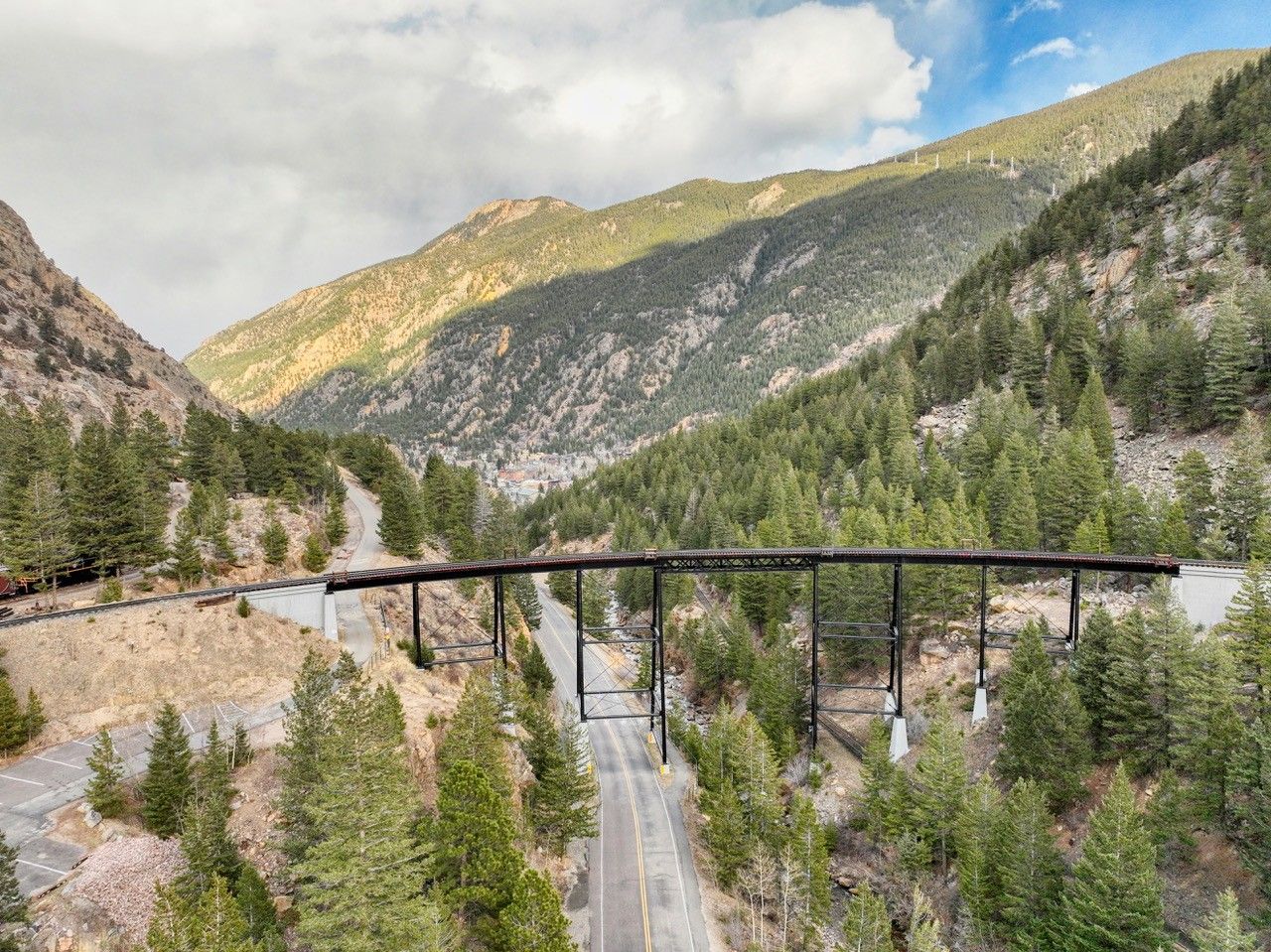 Mountain railway trestle bridge spanning a forested valley with rocky hills and a cloudy sky.