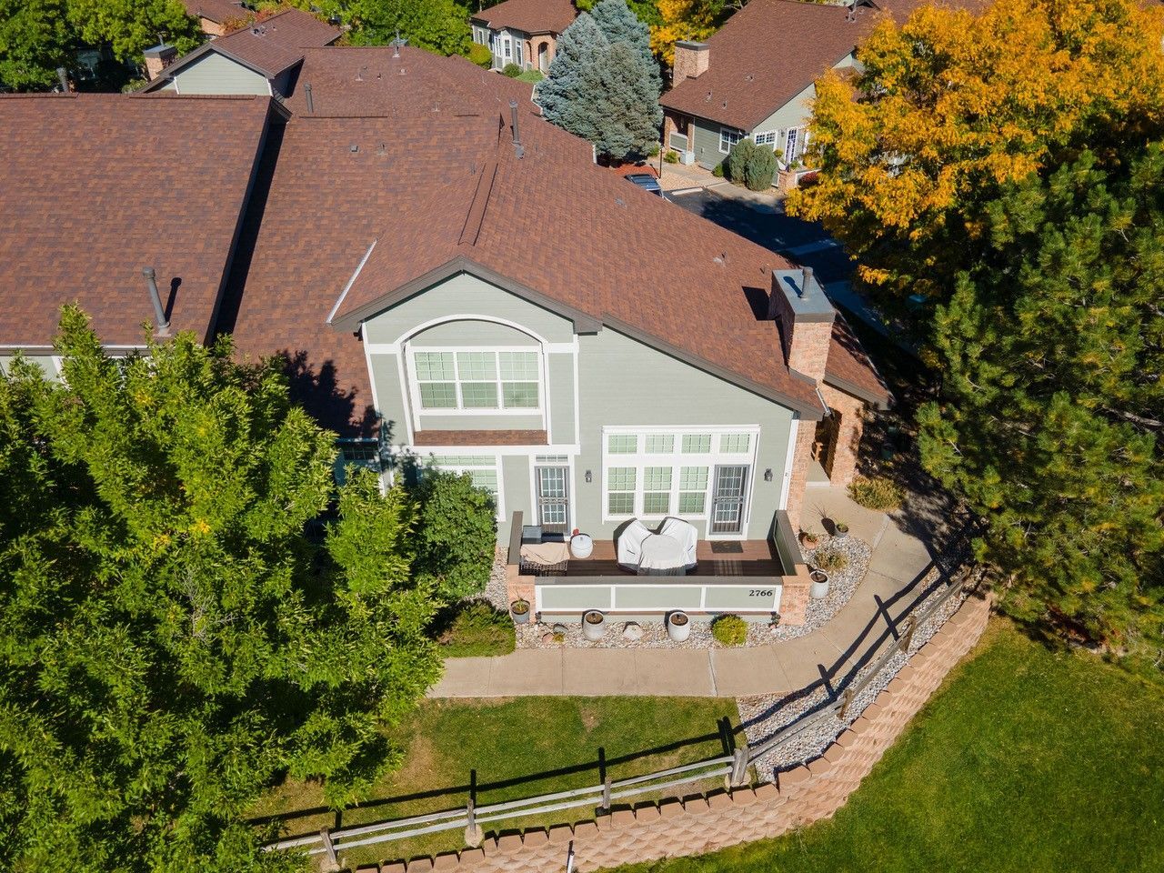 Aerial view of a light gray house with red roof, patio furniture, and a fenced backyard surrounded by trees.