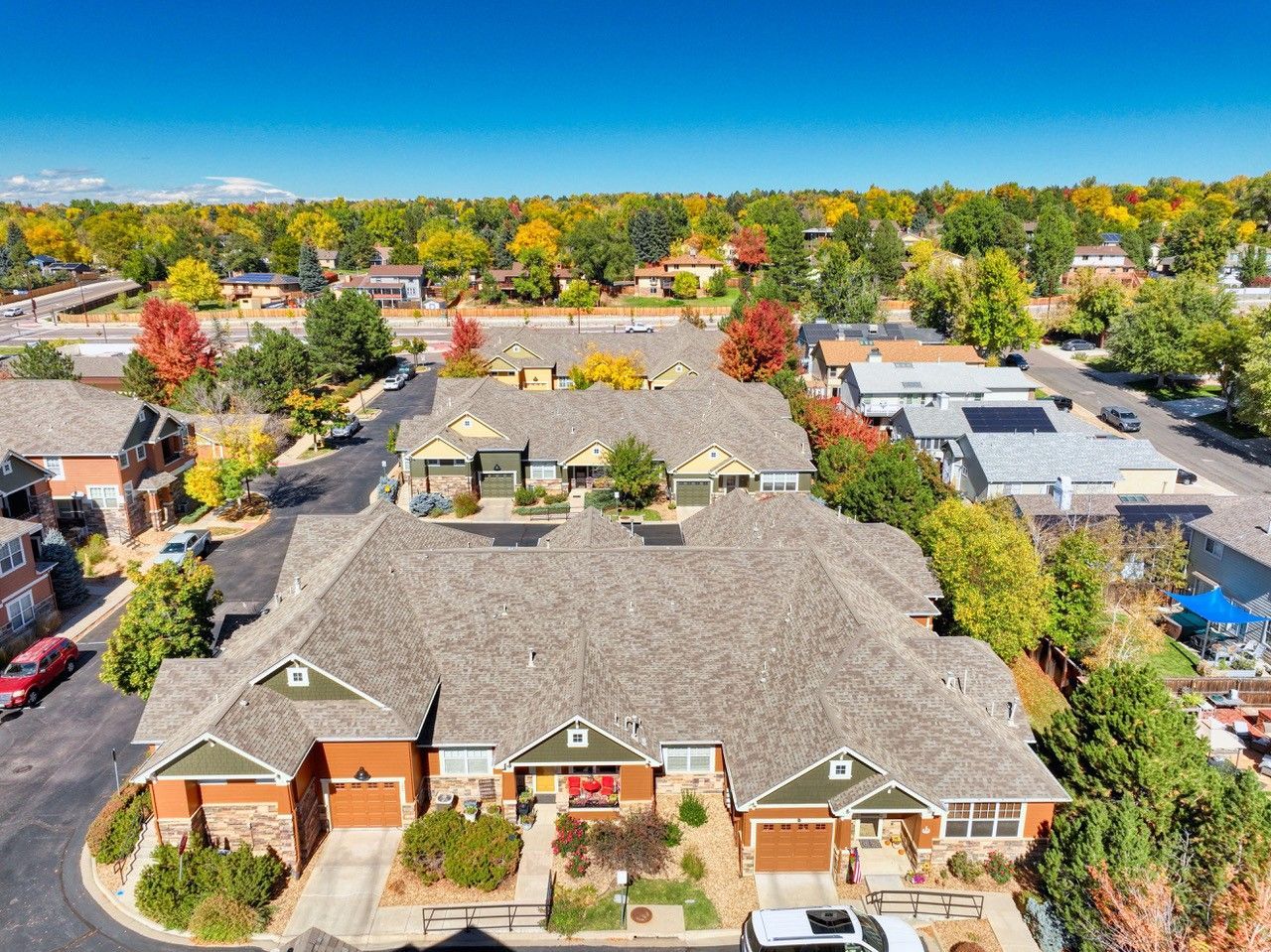 Aerial view of a suburban apartment complex with gray roofs, trees, and parked cars in a sunny neighborhood.