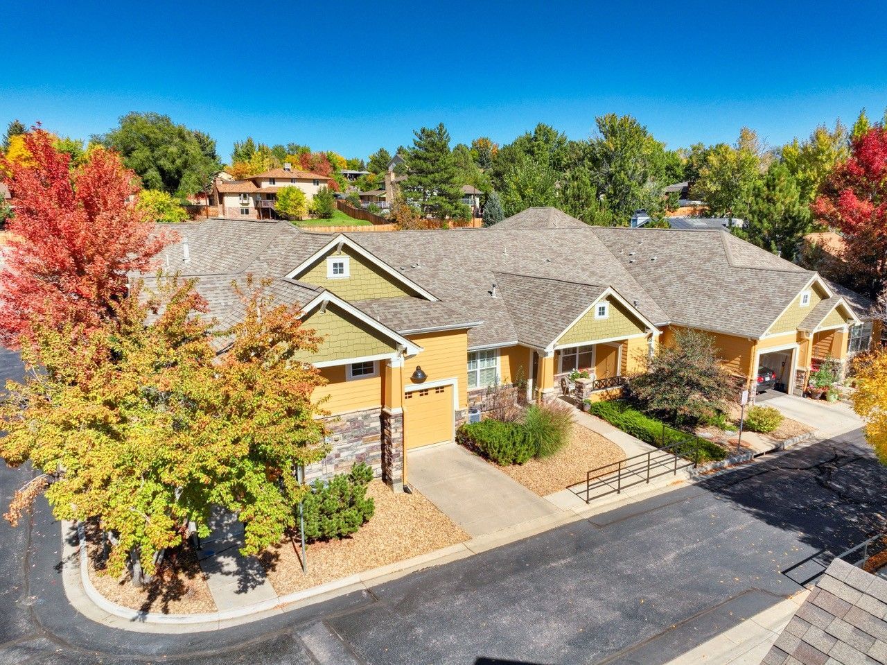 Aerial view of a row of tan townhomes with gray roofs and autumn-colored trees along a paved drive.