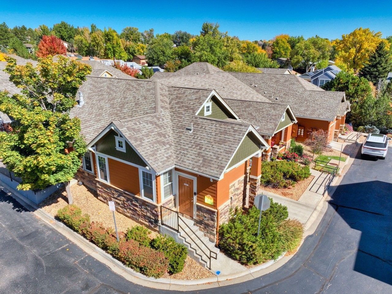 Aerial view of a brown house with a gray roof, front porch, and surrounding trees on a sunny street.