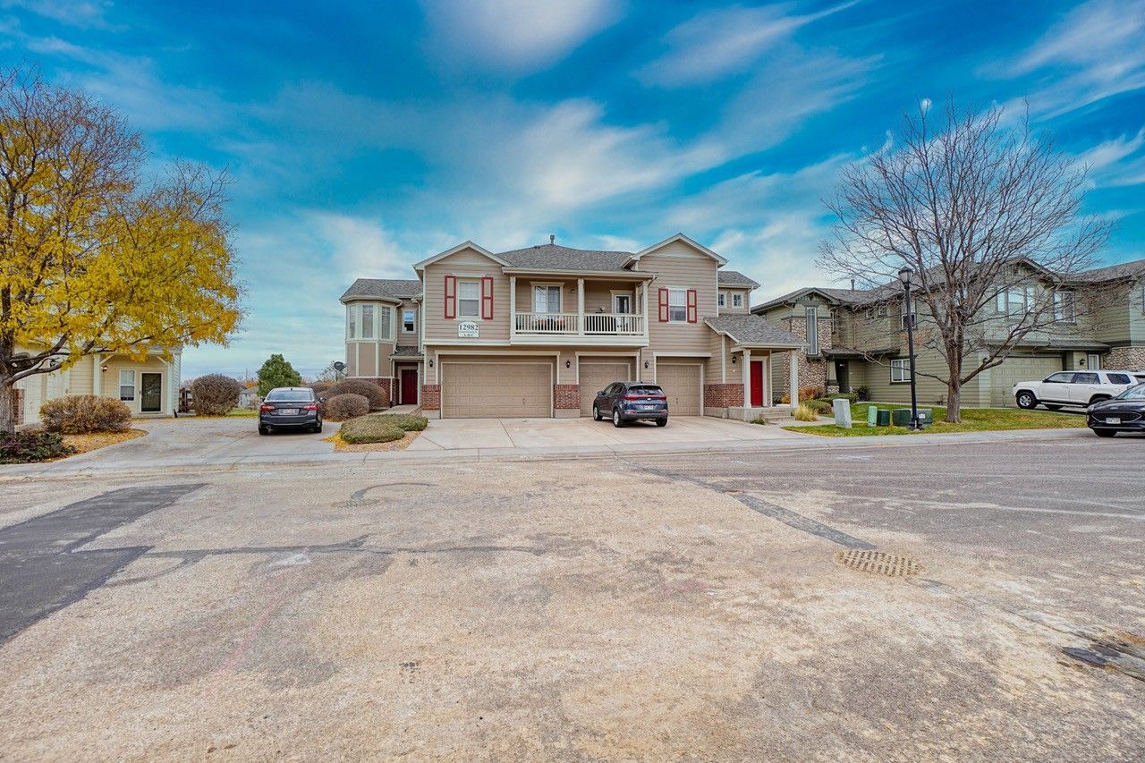 Two-story beige apartment building with balconies and parked cars in a gravel lot.