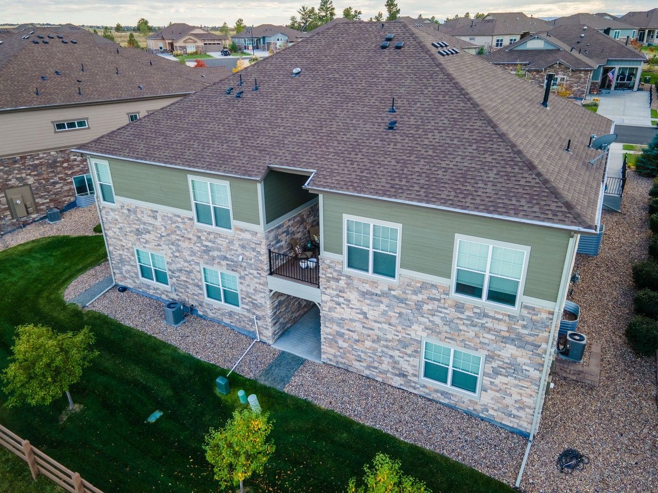 Aerial view of a two-story stone house with a brown roof and landscaped yard in a suburban neighborhood.