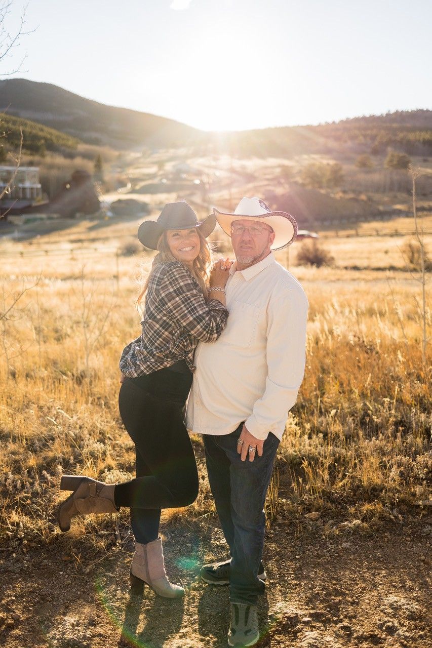 Couple in cowboy hats pose outdoors, smiling, with a golden sunset in the background.