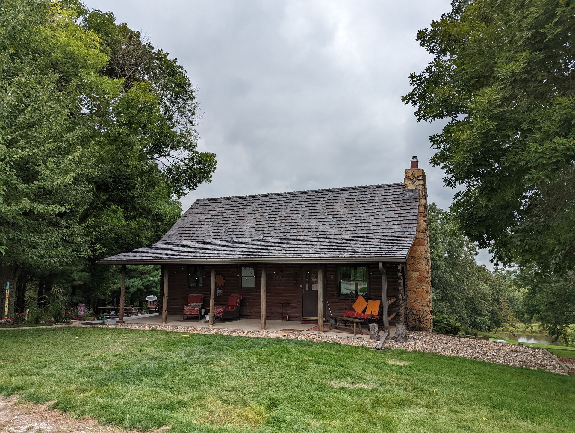 A log cabin with a porch and chairs is surrounded by trees on a cloudy day.