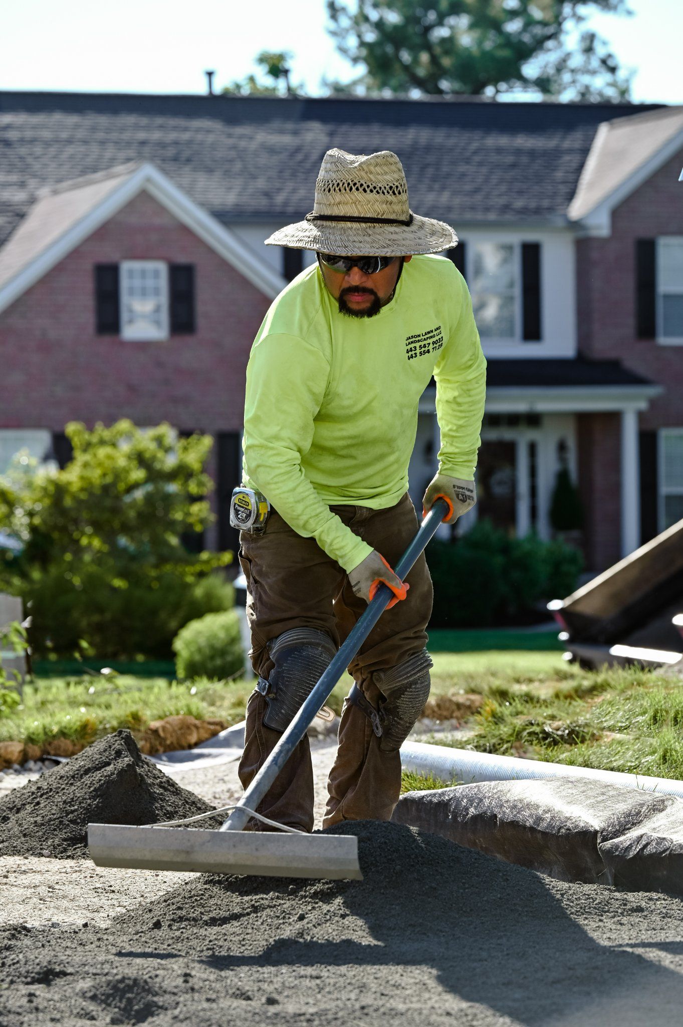 A man wearing a straw hat and sunglasses is raking concrete in front of a house.