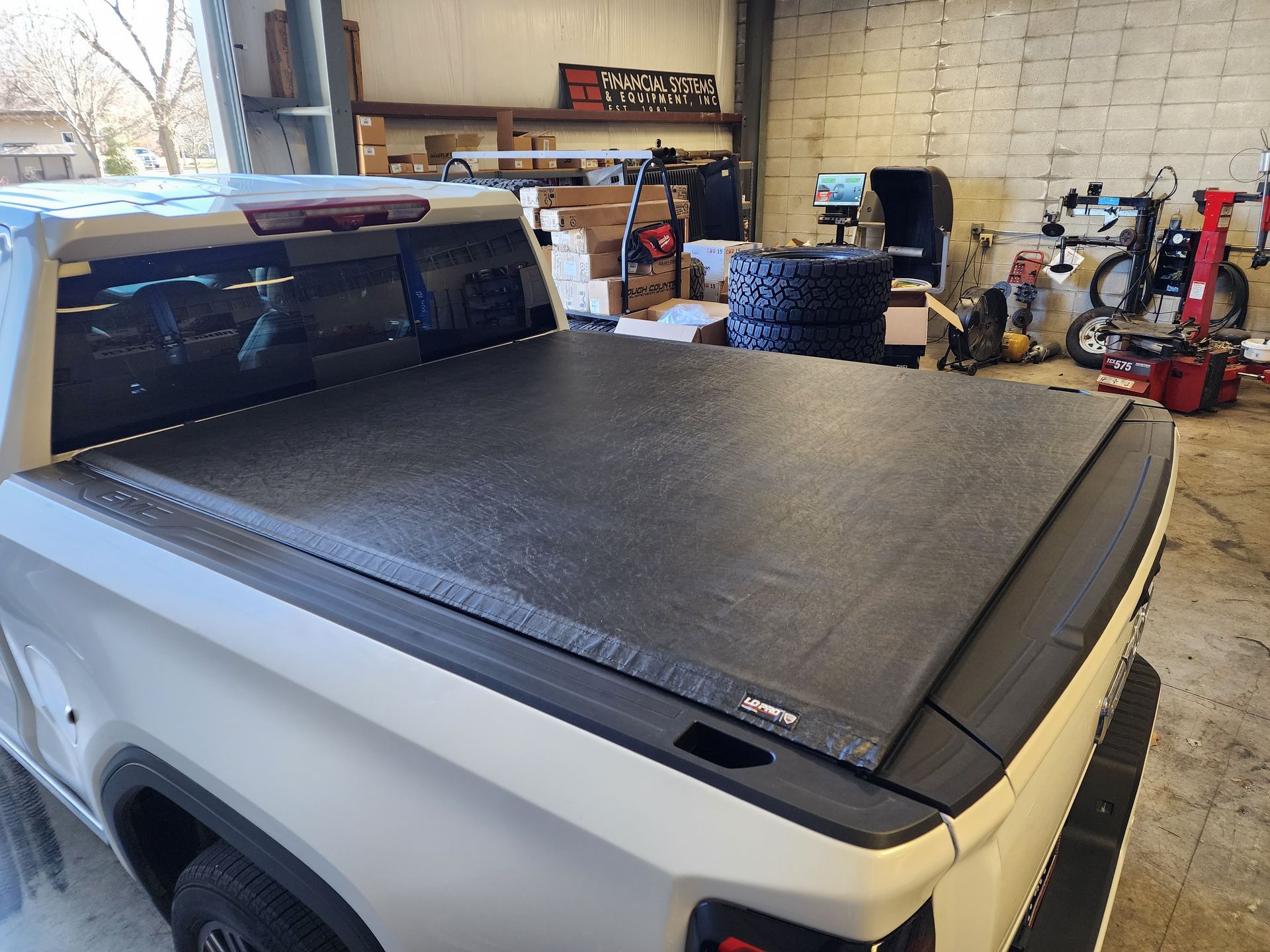 White pickup truck with a black tonneau cover parked in a garage with tools and spare tires.
