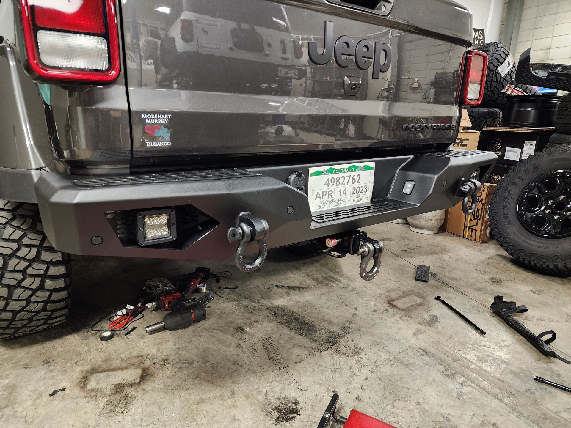 Jeep rear in garage with tow hitch, tools on floor, and a detached spare tire nearby