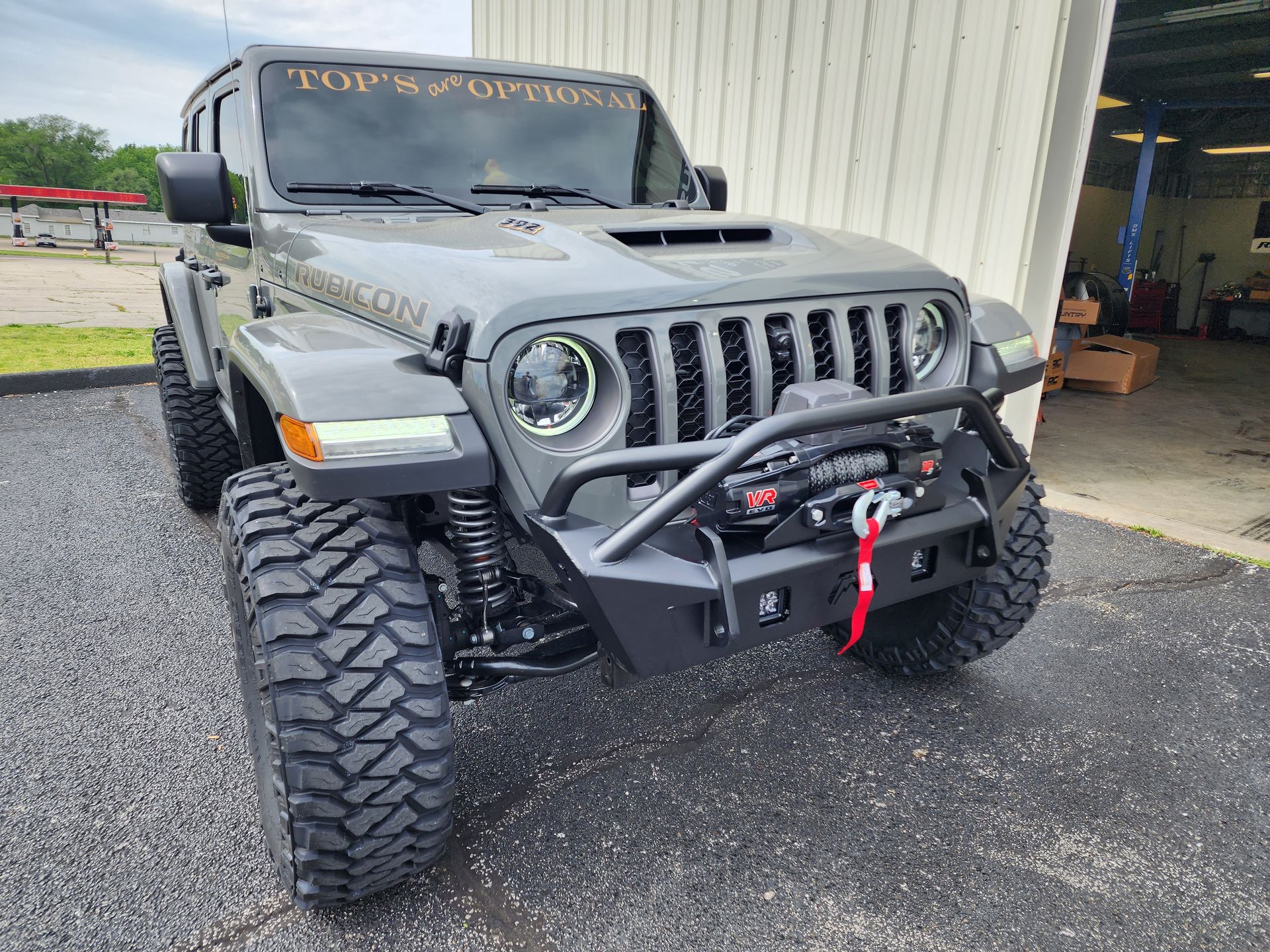 Gray Jeep Wrangler with off-road tires and front winch parked outside a garage.
