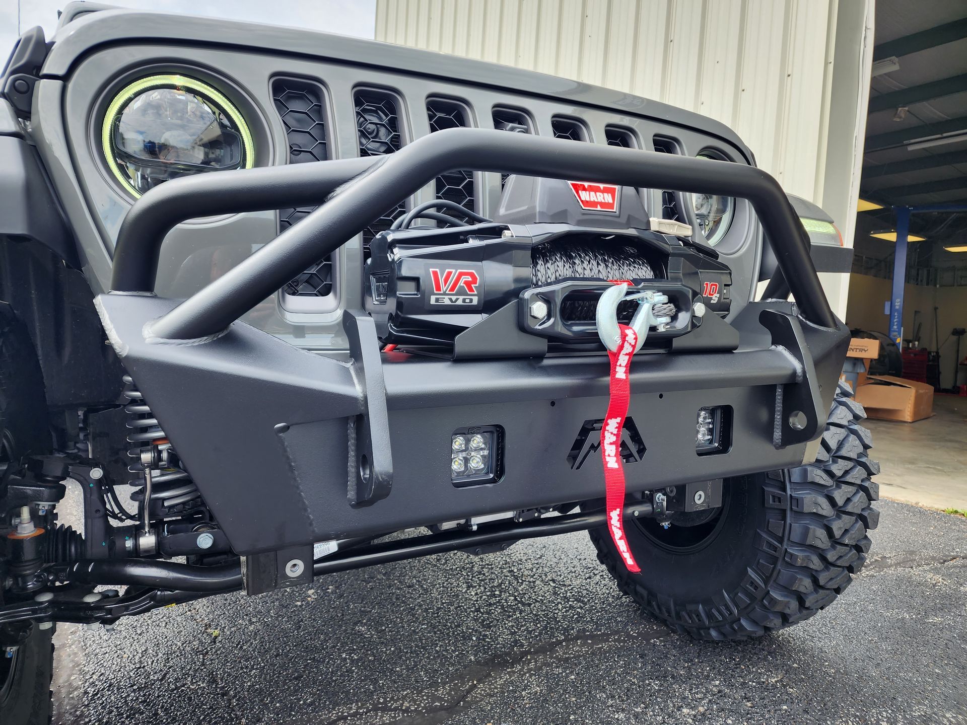 Grey off-road jeep front with winch and red tow strap, parked outside a garage