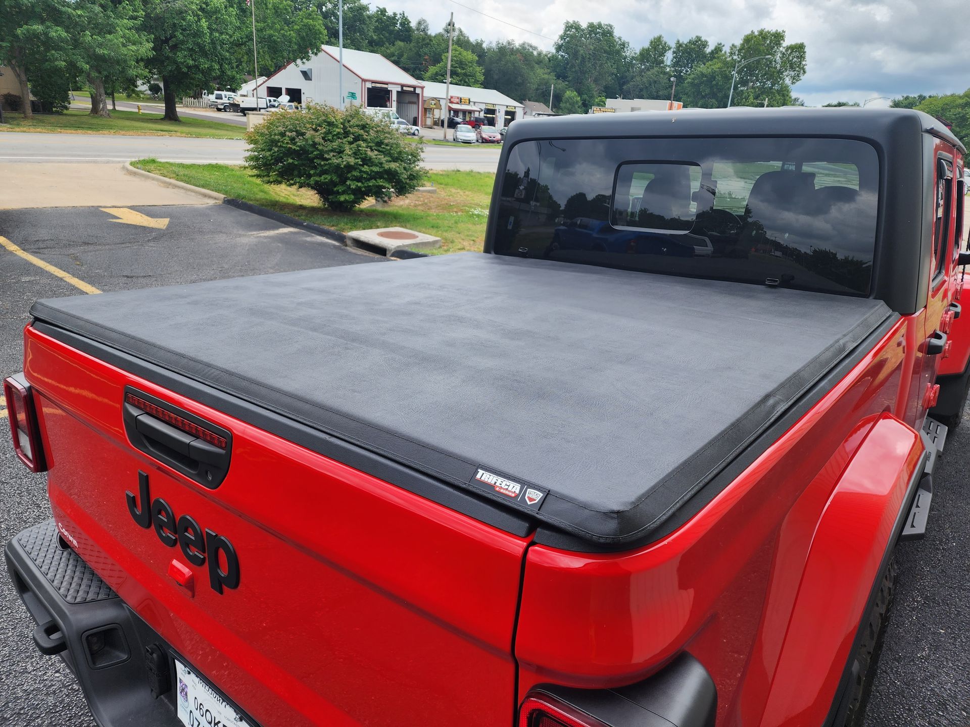 Red Jeep pickup with a black tonneau cover parked in a driveway near houses.