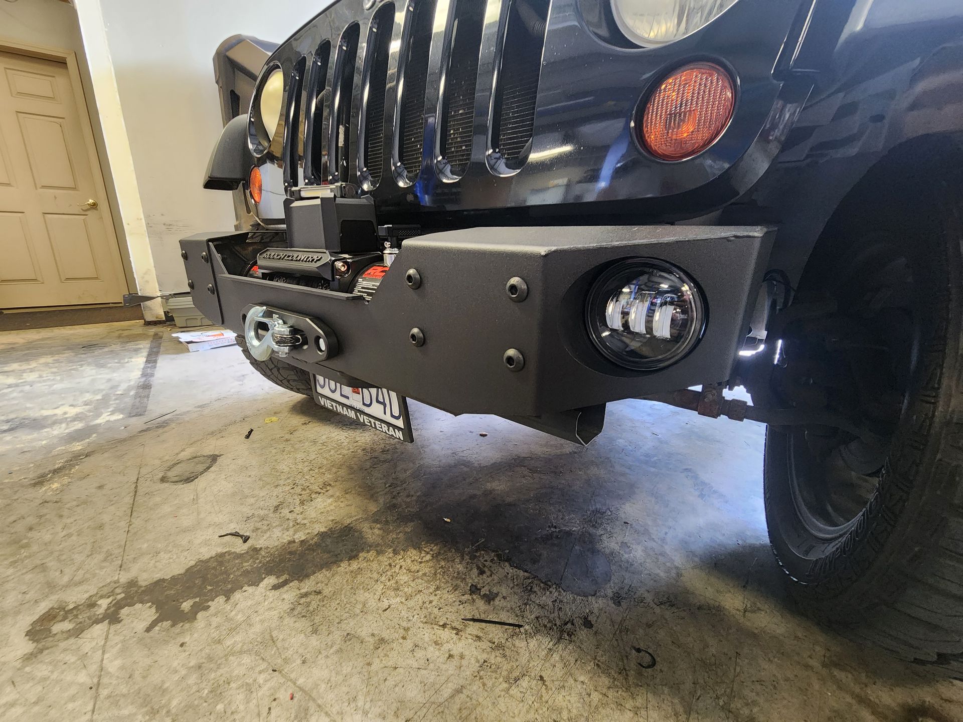 Black Jeep front bumper and grille parked in a garage, with off-road lights and a winch plate.