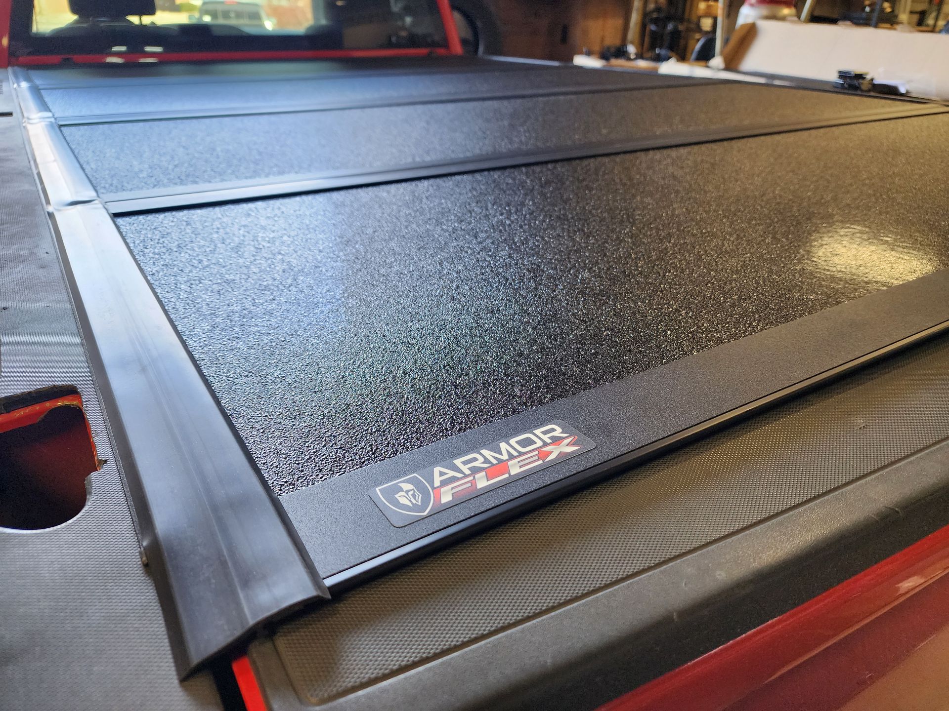 Black textured folding tonneau cover on a pickup truck bed, with AMOS logo visible.