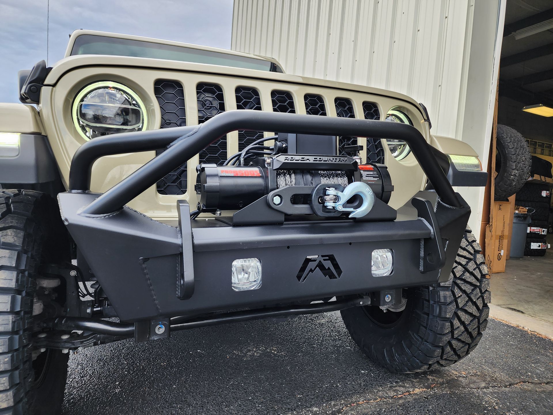 Modified beige Jeep with off-road bumper and winch parked outside a garage