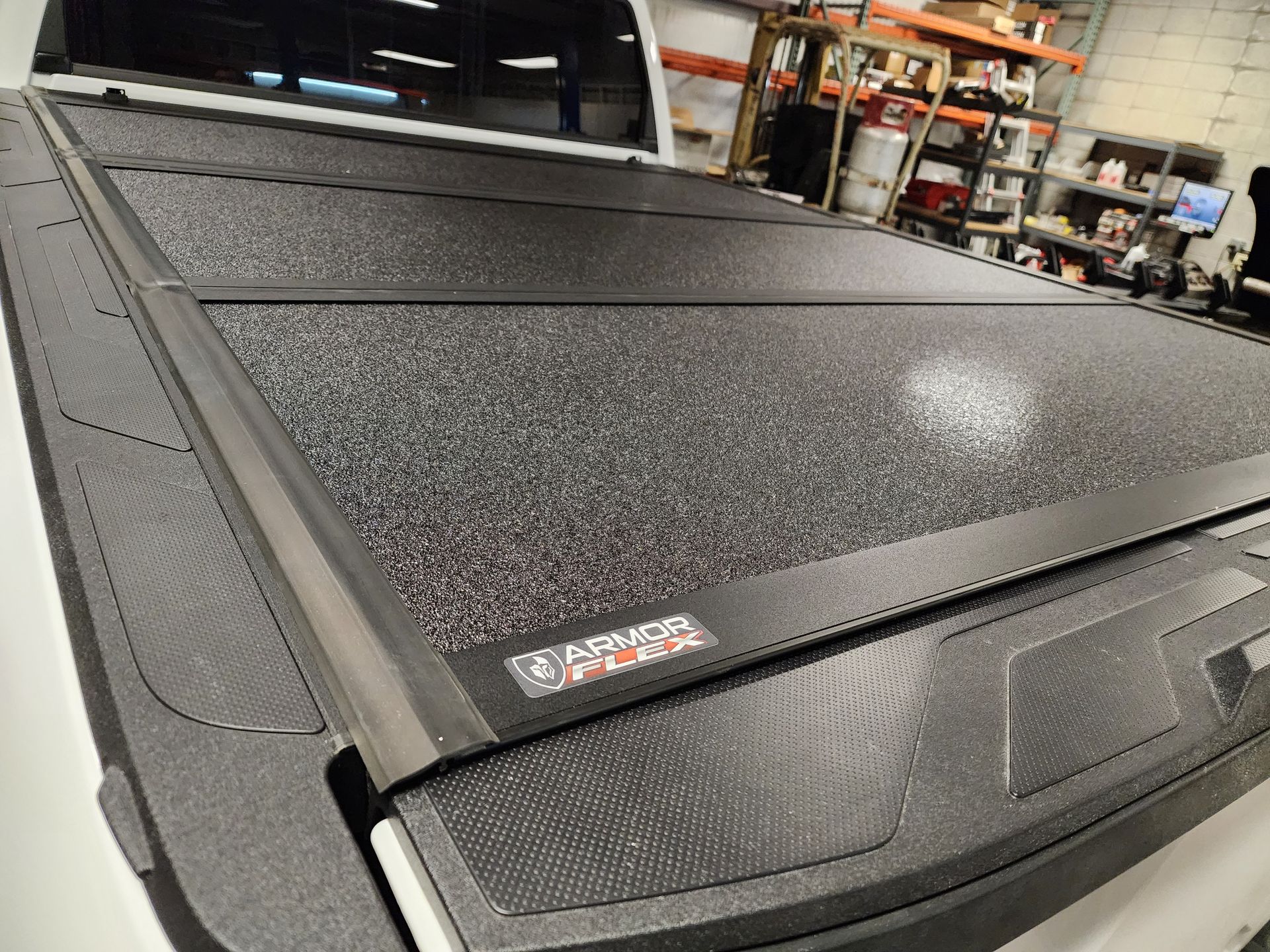 Black textured truck bed cover in a garage, shown close-up with rear window and shelves in the background