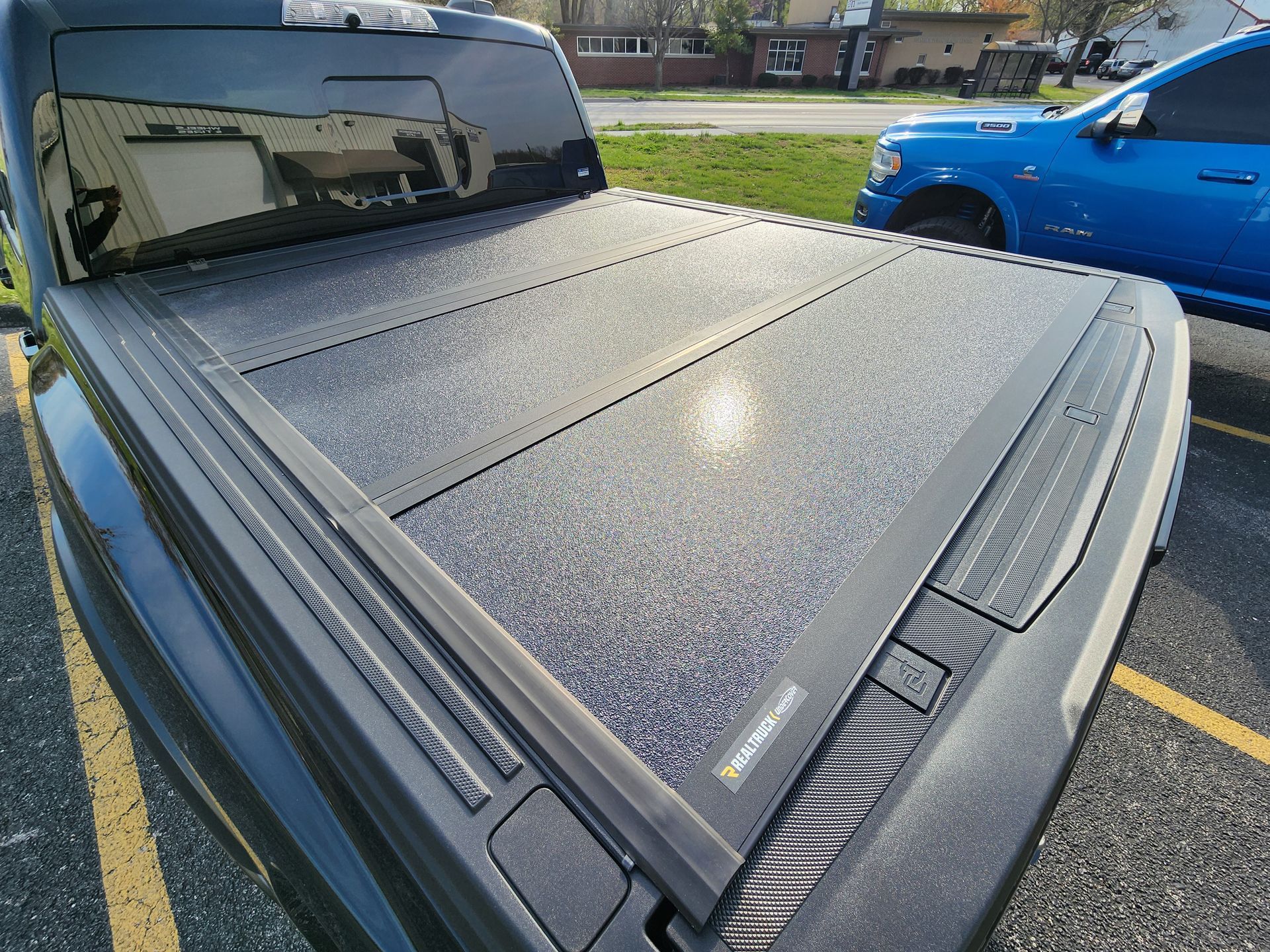 Close-up of a gray truck with a textured tonneau cover parked beside a blue pickup in a lot.