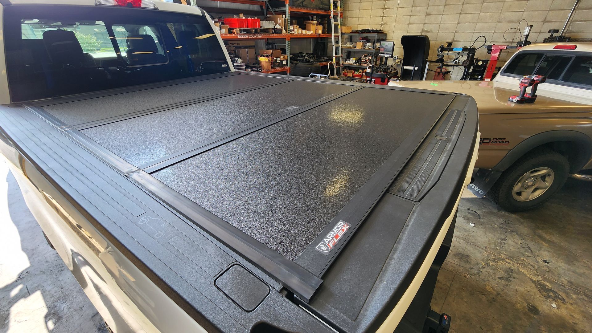 Black truck bed cover with water droplets in a garage, with another vehicle visible nearby