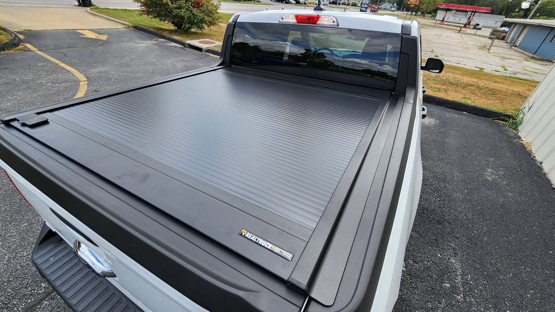 Black pickup truck bed covered with a textured tonneau cover, viewed from above on a driveway.