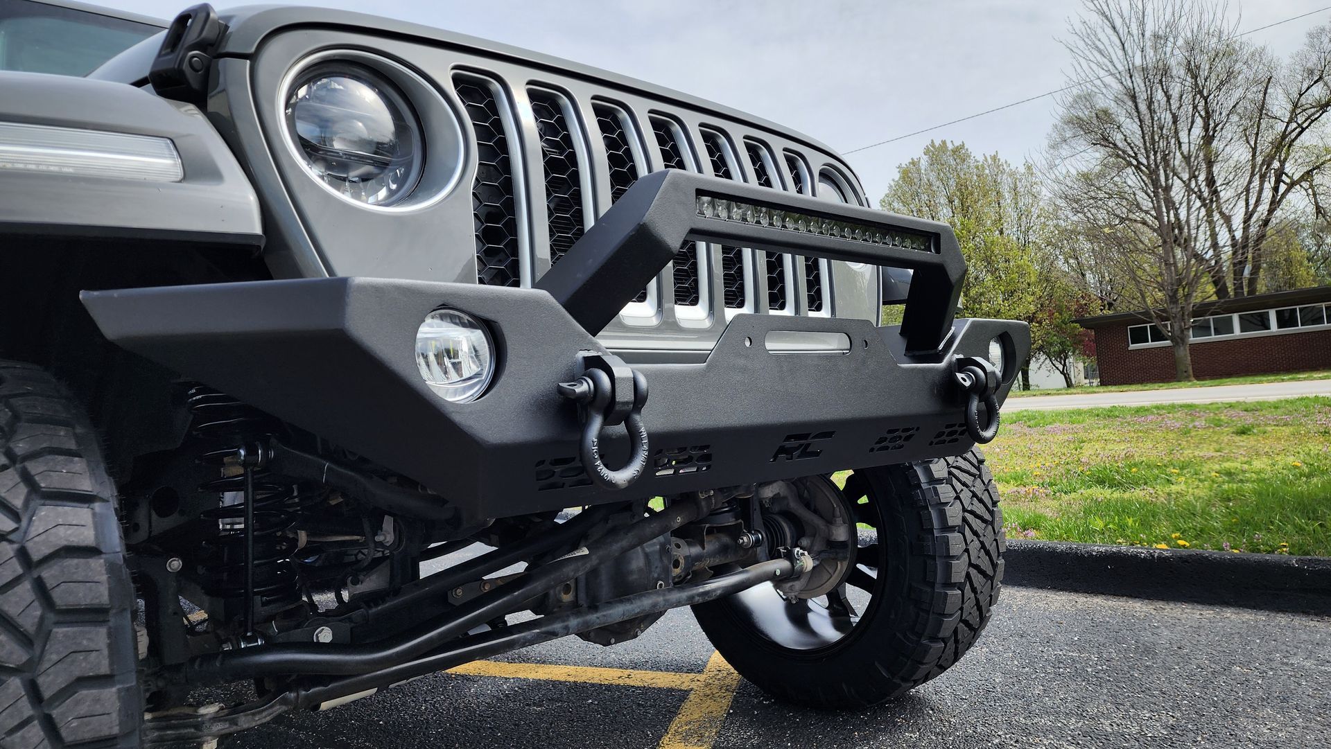 Gray Jeep Wrangler with off-road bumper and large tires parked on a driveway near grass and trees