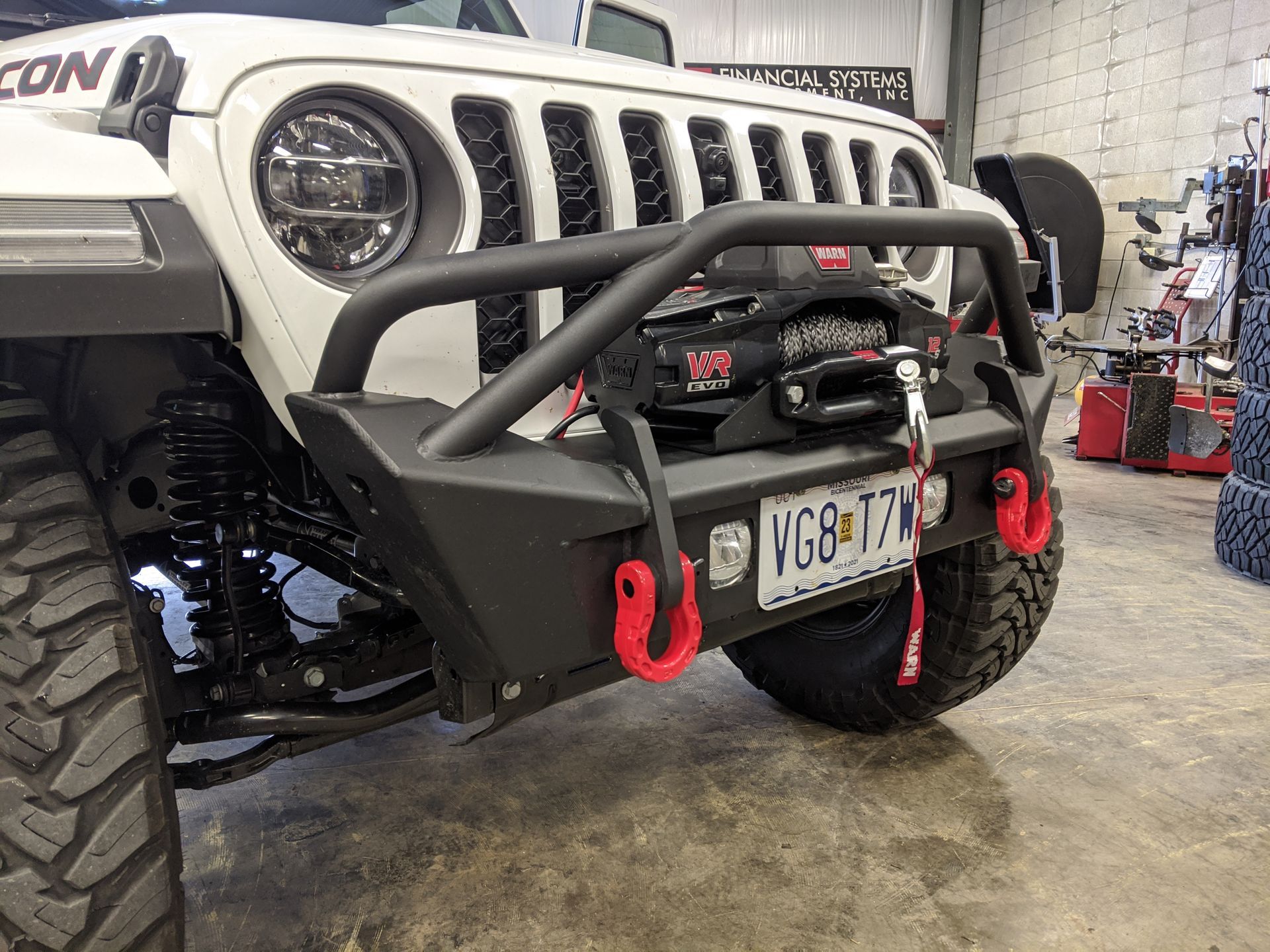 White off-road truck with black bull bar, red tow hooks, and winch in a garage.