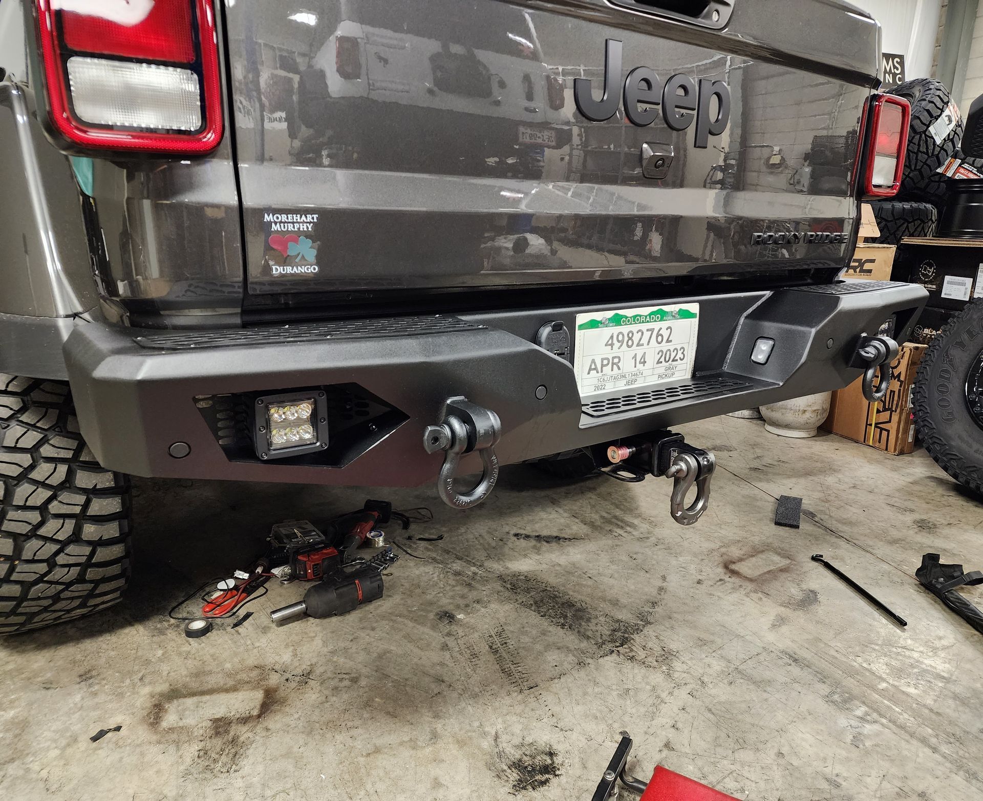 Rear view of a gray Jeep in a garage, showing a tow hitch and bumper accessories.